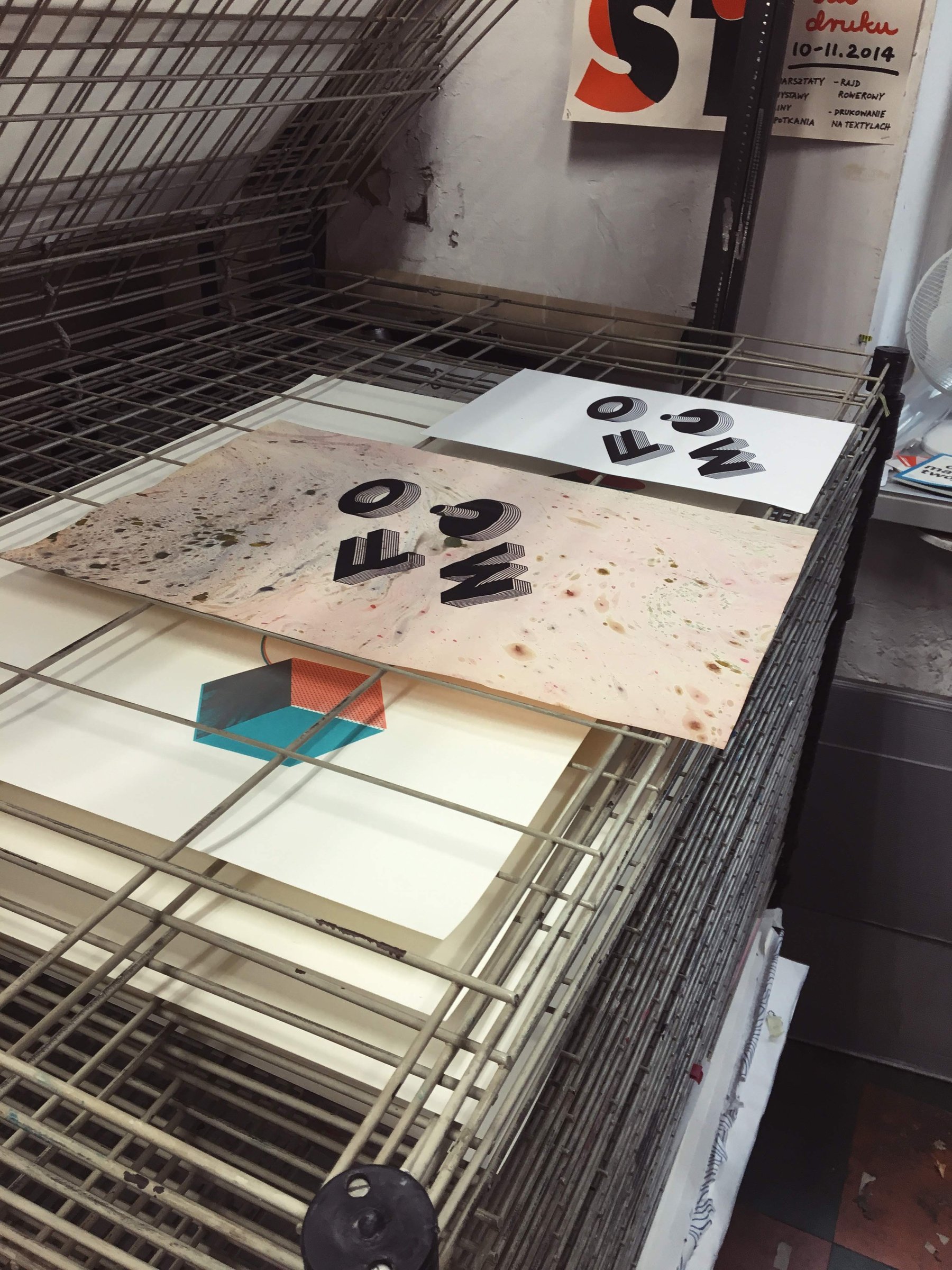 Two prints of the isometric FOMO drying on a wire rack, one on marbled pink paper and one on white.